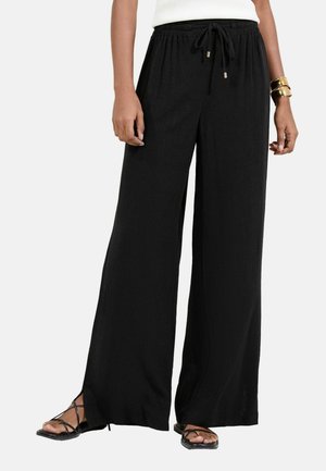 Woman wearing black wide-leg pants with elastic waist and drawstring, paired with black strappy sandals and gold bracelets.