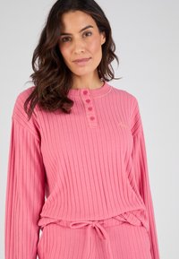 Top rosa de manga larga con textura de canalé, presenta un escote redondo, tapeta de cuatro botones y un sutil bordado. Textura suave y corte relajado.
