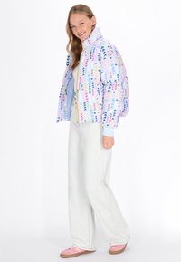 Jachetă pufoasă colorată pe un fundal albastru, decorată cu modele de săgeți în roz, galben și bleumarin. Are un fermoar galben și un guler înalt.