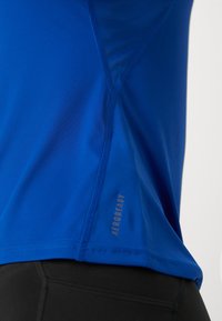 Närbild av en blå atletisk tröja med texten "AEROREADY" nära sidsömmen, bärs över svarta träningsleggings.