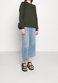 Olivově zelená mikina s volným střihem, v kombinaci s lehkými modrými denimmovými culotami. Černé platformové sandály mají pletený design pásků.