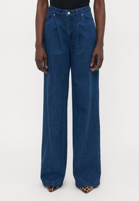 Oseba nosi temno modre široke hlače z visokim pasom in pregnami, črno majico ter špicaste čevlje z leopardjim vzorcem, stoječa ob nevtralni ozadju.