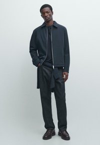 Ung mand stående med hænderne i lommerne iført en sort lynlåstrøje, mørke bukser og mørkebrune sko mod en ensfarvet baggrund.