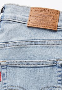 Jean en denim bleu clair avec une étiquette en cuir marron présentant un texte en relief. Couture visible le long des poches et de l'ourlet. Logo rouge à l'arrière.