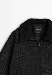 Veste noire texturée avec un col côtelé épais et une fermeture éclair devant, montrant l’étiquette de la marque « Massimo Dutti » à l’intérieur du col.