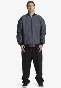 Denimbomberjacka med dragkedja fram, ribbade ärmslut och krage. Har en gummiloggopatch på bröstet. Matchas med svarta baggy-byxor.