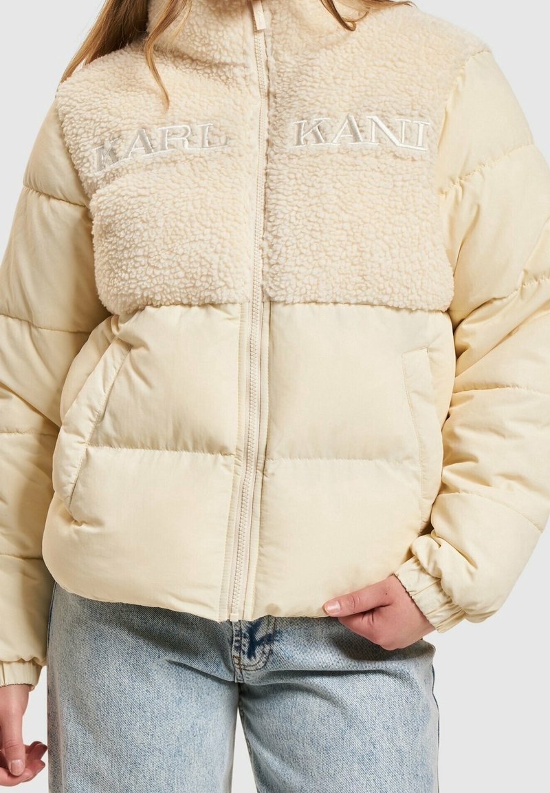 Doudoune beige avec panneau supérieur en sherpa et broderie "KARL KANI", portée avec un jean bleu clair, main dans la poche.