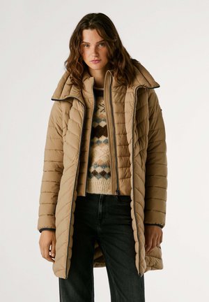 Giubbotto imbottito beige con silhouette lunga, texture trapuntata, colletto alto e zip. Indossato sopra un maglione a fantasia e jeans neri.