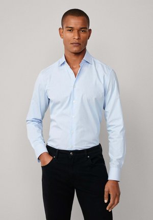 Mann in hellblauem Hemd mit Knopfleiste und schwarzen Hosen, steht mit einer Hand in der Tasche vor einem schlichten Hintergrund.