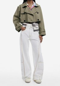 Trinchera recortada verde oliva con botones dobles sobre una camisa estampada; combinada con jeans de pierna ancha blancos con costuras contrastantes.