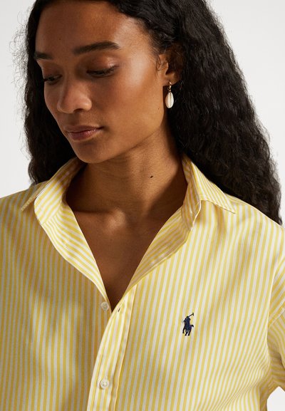 Sieviete ar gariem cirtainiem matiem, valkājot dzeltenu un baltu svītrainu kreklu ar tumši zilu logo un vienu gliemežvāka auskaru, skatoties uz leju.