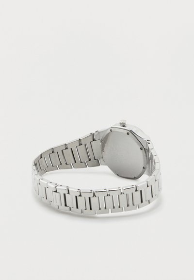 Ασημένιο μεταλλικό ρολόι χειρός με συνδεδεμένη λωρίδα, εμφανιζόμενο από πίσω, ακουμπισμένο σε φωτεινή επιφάνεια με εμφανή το στέμμα και τις βίδες της κάσας.