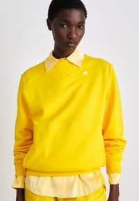 Felpa gialla con polsini e orlo a coste, indossata sopra una camicia a righe gialla chiaro. Presenta un piccolo logo sul lato sinistro del petto.
