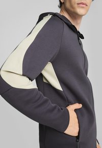 Sudadera con capucha en gris oscuro con acentos en crema texturizados en las mangas. Cuenta con un bolsillo con cremallera y un acabado de tela suave.
