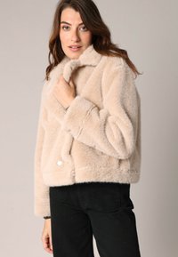 Corta chaqueta de piel sintética en beige claro con una textura suave, cuello ancho y cierre de botón en la parte delantera, combinada con pantalones negros.