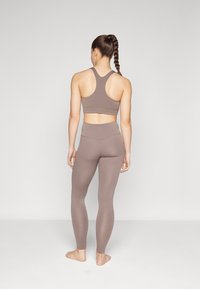 Ruskea urheil bra ja korkeavyötäröiset leggings, tiukka muotoilu, sileä pinta ja minimaalinen saumoja. Malli nähtynä takaa, paljain jaloin.