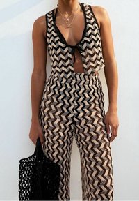 Pletený top a odpovídající kalhoty v béžovém a černém zigzag vzoru, s krátkým střihem a černou síťovou taškou v ruce.