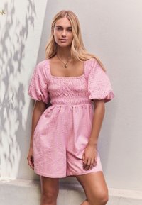 Vaaleanpunainen haalari, jossa on puffy-hihat, ryppyinen vartalo ja pystysuuntaiset kuvioidut raidat. Valmistettu kevyestä kankaasta, sopii lämpimään säähän.