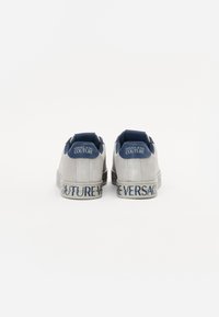 Sapatilhas de camurça cinza com detalhes azuis, apresentando um colarinho acolchoado e sola com marcação "VERSACE" em texto azul. Textura suave, design de perfil baixo.