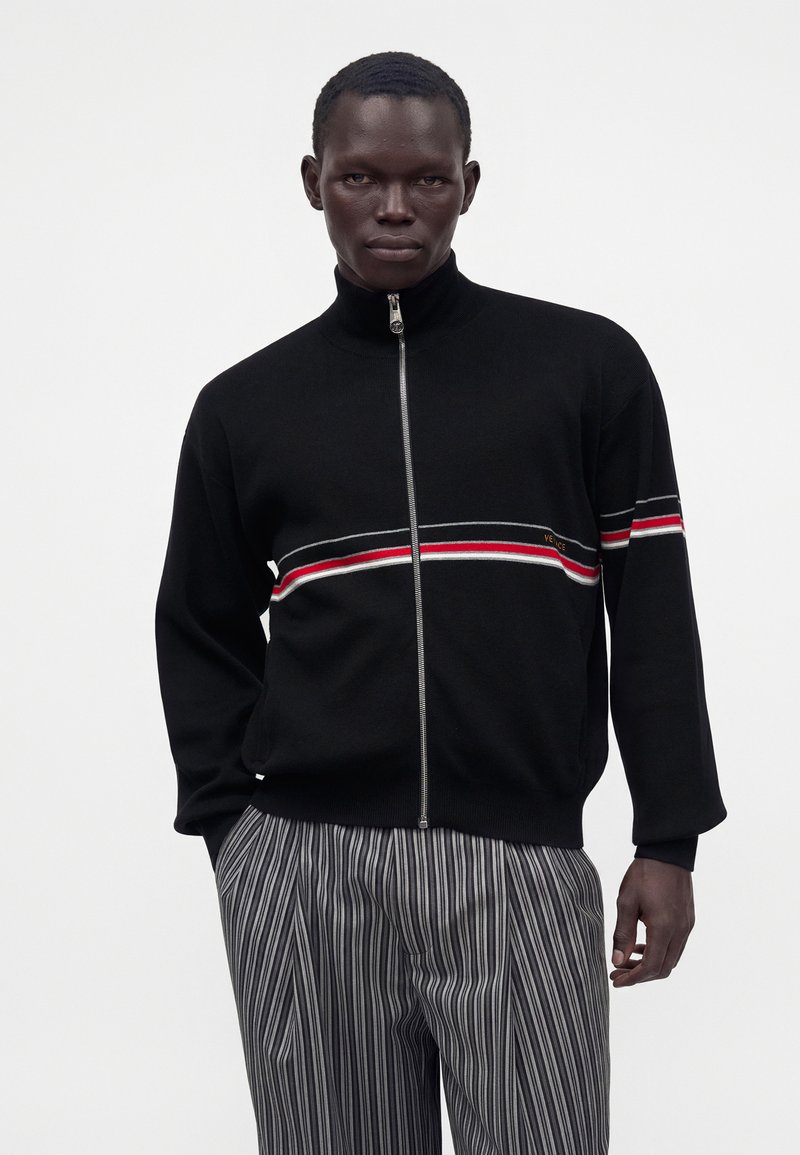 Homme portant un pull zippé noir avec des rayures rouges et blanches et un pantalon rayé gris, debout devant un fond blanc uni.