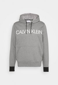 Sweat à capuche gris avec un grand logo blanc "CALVIN KLEIN", cordons de serrage noirs et une doublure intérieure à motifs. Présente des poignets et une taille côtelés.