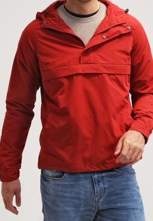 Man wearing a red hooded anorak jacket over a beige sweater and blue jeans, shown from mid-chest to hips.