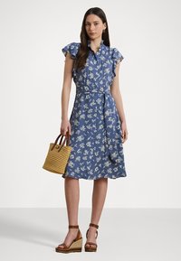 Blue floral dress with ruffled sleeves, cinched waist, and belt. Model holds a woven straw bag and wears brown wedge sandals.