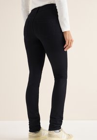 Jeans ajustados de mezclilla negra con una cintura de media altura, que cuentan con bolsillos traseros y una textura suave, combinados con zapatillas crema.