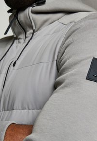 Giacca imbottita grigio chiaro con tessuto testurizzato sulle maniche, cappuccio regolabile con cordini e una patch nera con logo sul braccio.