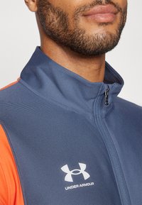 Sportjacke mit Reißverschluss in Marineblau und orangefarbenen Akzenten. Mit gerippter Textur und weißem Under Armour-Logo auf der Brust.