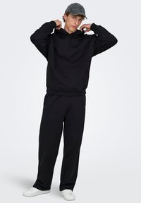 Schwarzer Hoodie mit Kängurutasche, kombiniert mit passenden schwarzen Jogginghosen. Das Model trägt eine graue Kappe und weiße Turnschuhe. Schlichte Textur, lässiger Stil.