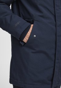 Navy blue jacket made of textured fabric, featuring a pocket, ribbed cuffs, and a snap button detail on the front.