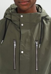 Veste à capuche vert olive en matière résistante à l'eau, dotée de deux poches zippées et de cordons noirs. Boutons-pression argentés visibles.