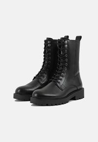 Zwarte leren combatboots hebben een hoge schacht, ronde neus en een stevige, geribbelde zool. De veters lopen omhoog aan de voorkant met metalen oogjes.