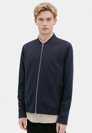 Ung mand med kort blondt hår iført en marineblå lynlåstrøje over en beige skjorte, stående foran en ensfarvet hvid baggrund.