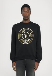 Črn sweatshirt z okroglo, sijočo zlato logotipom, ki vsebuje "VERSACE JEANS COUTURE." Rebrasti manšeti in spodnji rob, mehka tekstura, standardna prileganje.