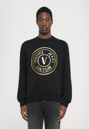 Svart sweatshirt med en rund, glänsande guldig logotyp som visar "VERSACE JEANS COUTURE." Ribbstickade muddar och fåll, mjuk textur, normal passform.