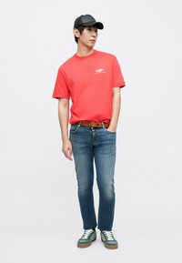 T-shirt en coton rouge avec un motif de poisson, jean en denim bleu, ceinture en cuir marron, et baskets vertes avec des lacets blancs ; portant une casquette noire.