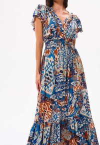 Monivärinen maxi-mekko, jossa on sekoitus sinisiä, ruskeita ja kreeminvärisiä kuvioita. Rypytetty hihansuu ja smockattu vyötärö, jossa on kerroksellinen hame.