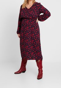 Vestido navy con estampado floral rojo, escote en forma de V, mangas largas y cintura elástica. Combinado con botas de vaquero rojas y una silueta relajada.