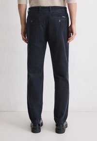 Marineblaue Cordhosen mit vertikalen Rippungen, zwei Gesäßtaschen und dezenten Branding-Details. Schmal zulaufendes Bein-Design und getragen mit schwarzen Schuhen.