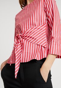 Blusa a righe rosse e bianche con maniche a tre quarti, caratterizzata da un dettaglio annodato sul davanti e una vestibilità ampia, abbinata a pantaloni neri.