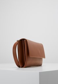 Ruskea nahkaboksi, jossa on läppäkiinnitys, sileä pinta, huomaamaton ompelu ja sivukahva kantamiseen. Yksinkertainen, suorakulmainen muotoilu.