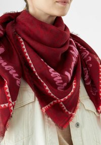 Femme portant une écharpe à motifs rouge profond avec des designs roses et blancs sur une veste beige clair avec des poches visibles et des boutons.