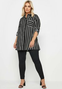 Maglietta a strisce bianche e nere con maniche lunghe e chiusura con bottoni, dotata di una tasca sul petto e un orlo posteriore leggermente più lungo. Indossata con pantaloni neri.