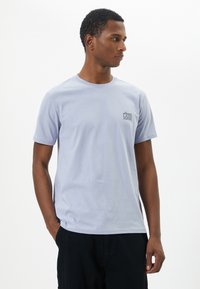 Camiseta de algodón morada con cuello redondo, mangas cortas y detalle de bolsillo gráfico en el frontal. Textura suave y corte relajado.