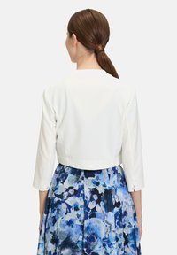 Witte cropped jas met driekwartmouwen, een gladde textuur en geen zichtbare sluitingen, gecombineerd met een blauwe bloemenrok.