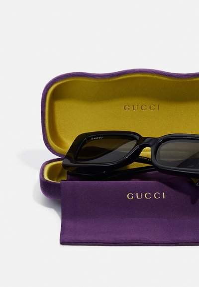 Lunettes de soleil noires dans un étui en velours violet avec un intérieur doré. Comprend un chiffon de nettoyage assorti en violet avec du texte doré.