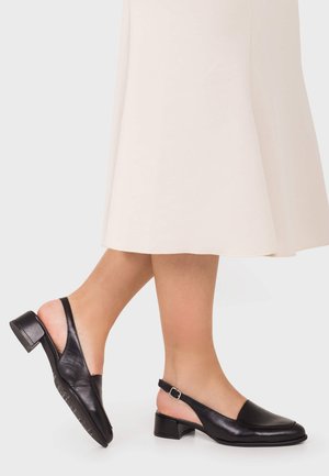 Persona con una falda midi color crema y zapatos negros slingback de tacón bajo, de pie con un pie ligeramente levantado sobre un fondo liso y claro.