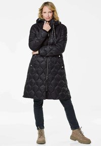 Zwarte gewatteerde pufferjas met een capuchon, voorzien van een rits aan de voorkant en een trekkoord, gecombineerd met donkere jeans en tan kleurige veterschoenen.